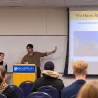 man standing behind podium gesturing to a projector and images of forests, with text that reads "we have always been here"
